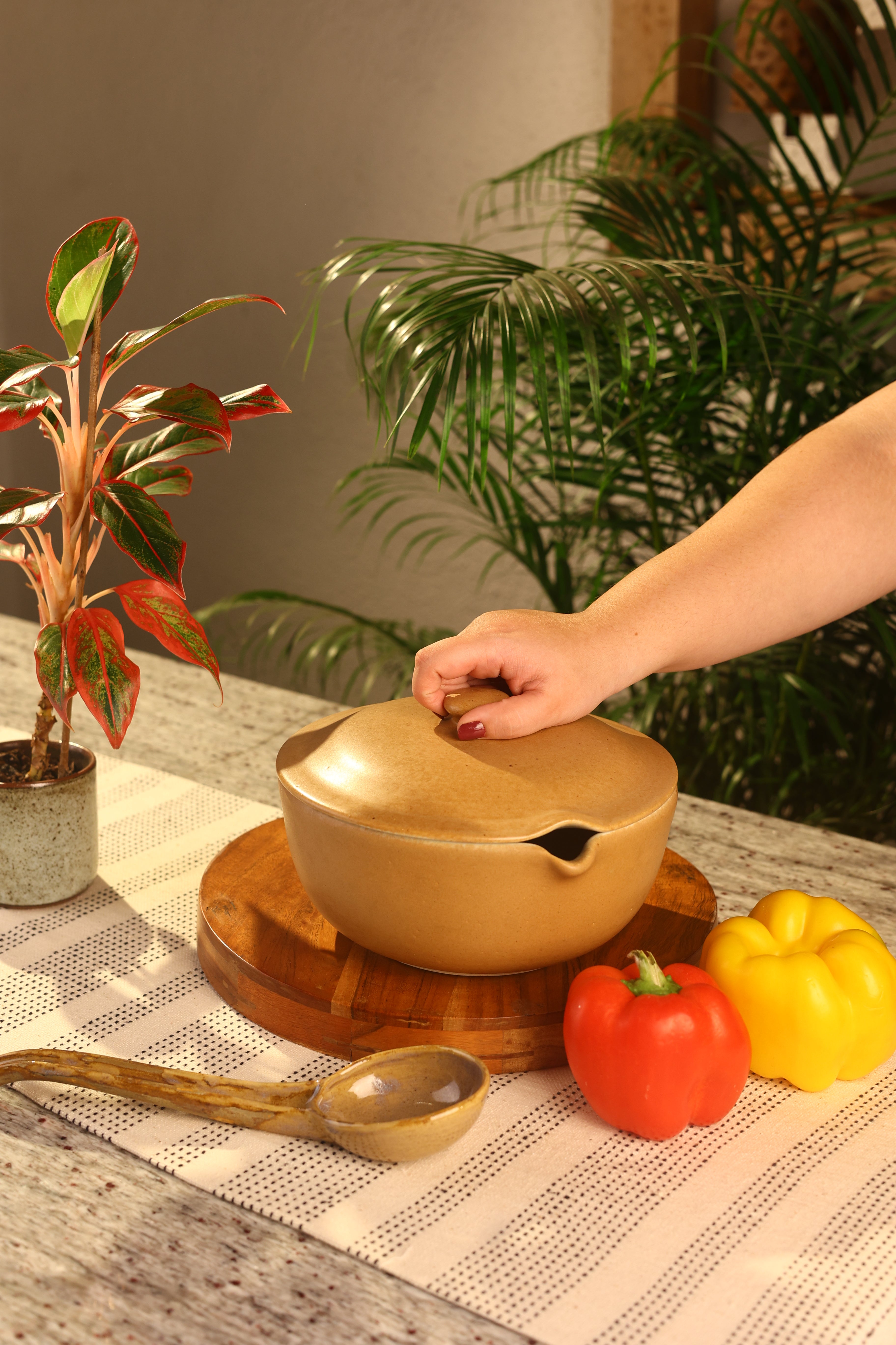 Person holding a mustard pot with a plant and colorful peppers in the background artisanal serveware India, handcrafted plates and bowls