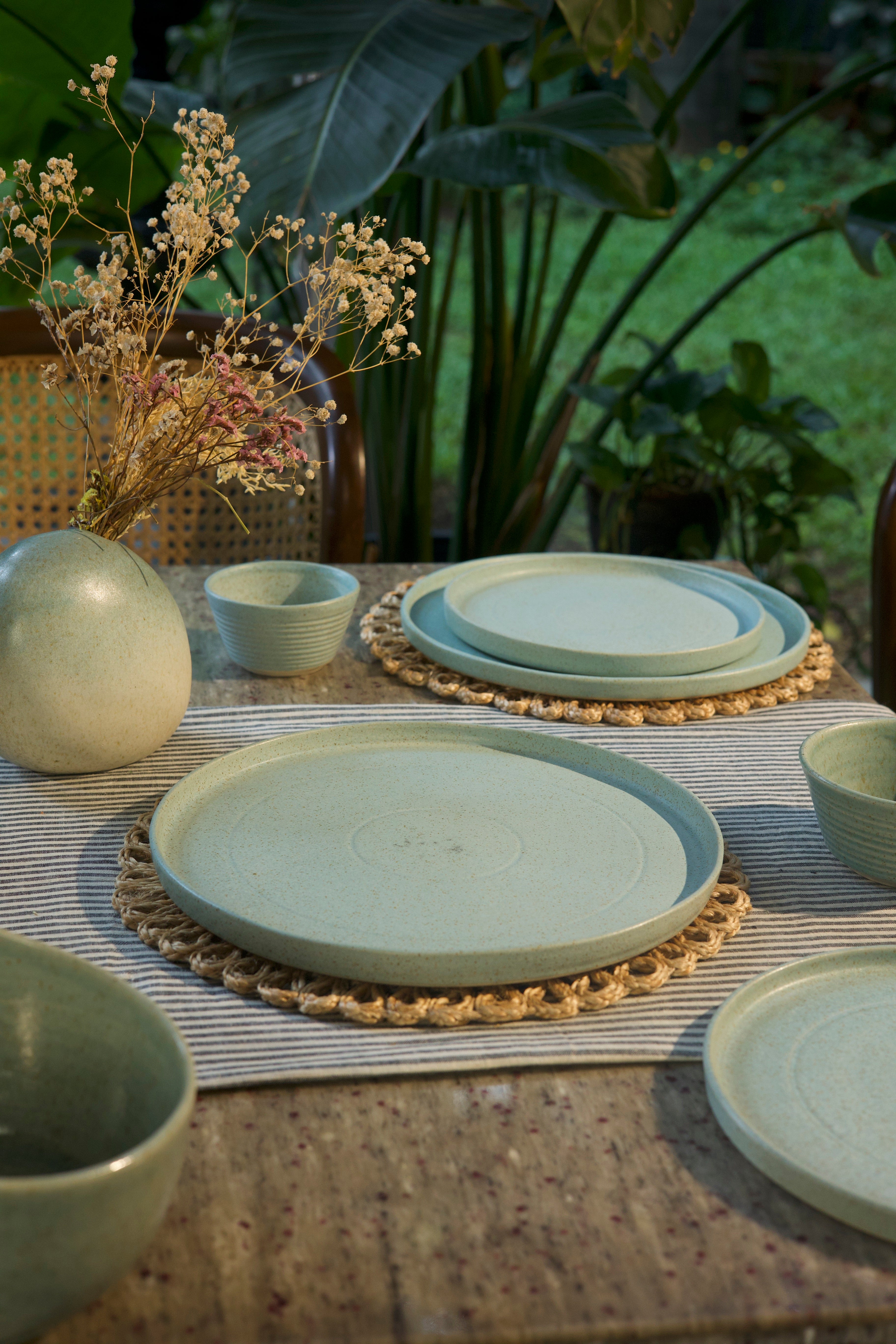 Set of ceramic plates and bowls on a table with a woven placemat, surrounded by plants. ceramic tableware India, handcrafted plates and bowls