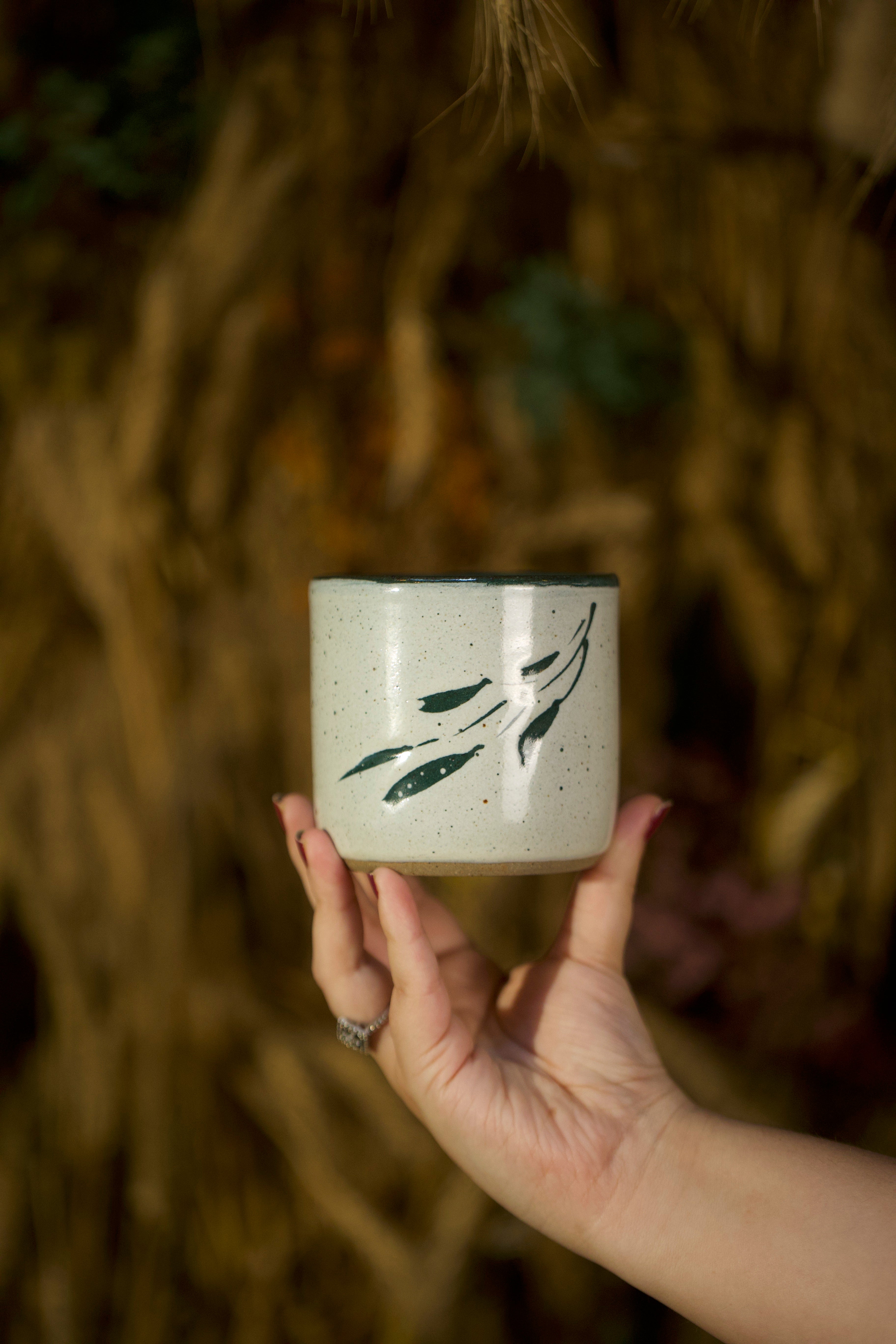 Hand holding a small ceramic cup with green leaf design against a blurred natural background, artisanal serveware India, handmade ceramics India