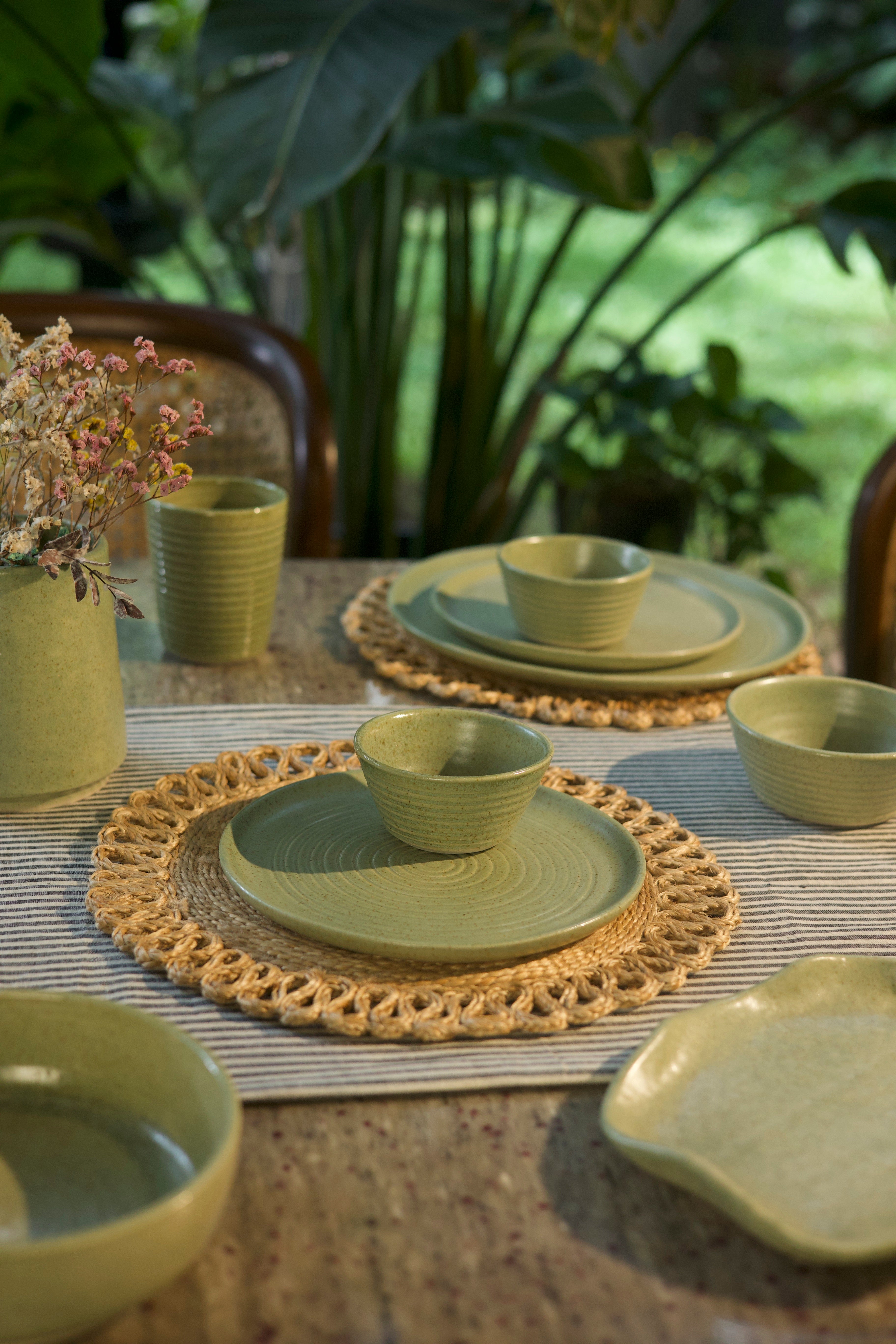 Green ceramic tableware set on a textured surface with plants in the background handmade dinnerware India, handcrafted plates and bowls