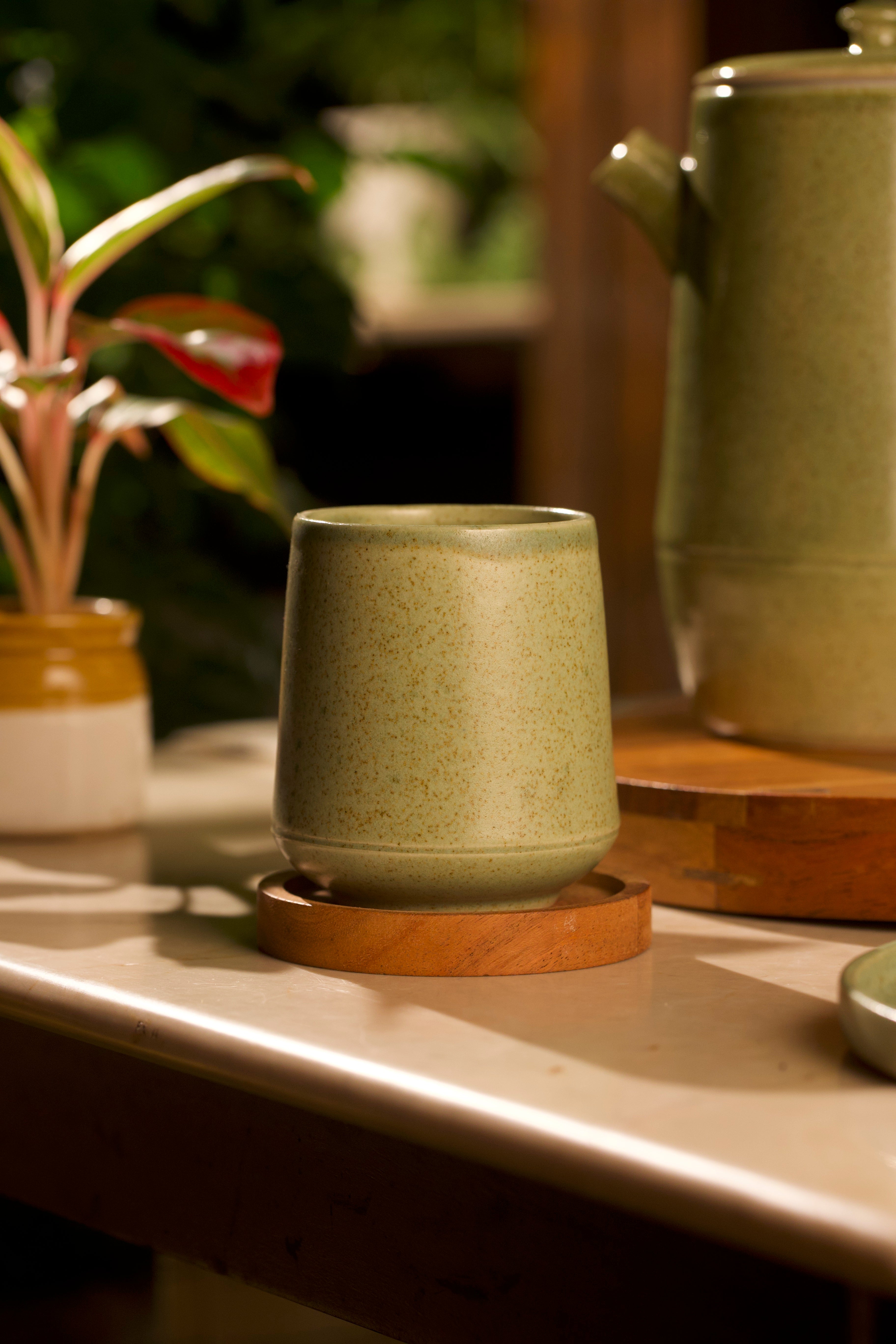 Green ceramic cup on a wooden coaster with a blurred background of plants and a teapot. artisanal serveware India, handmade ceramics India