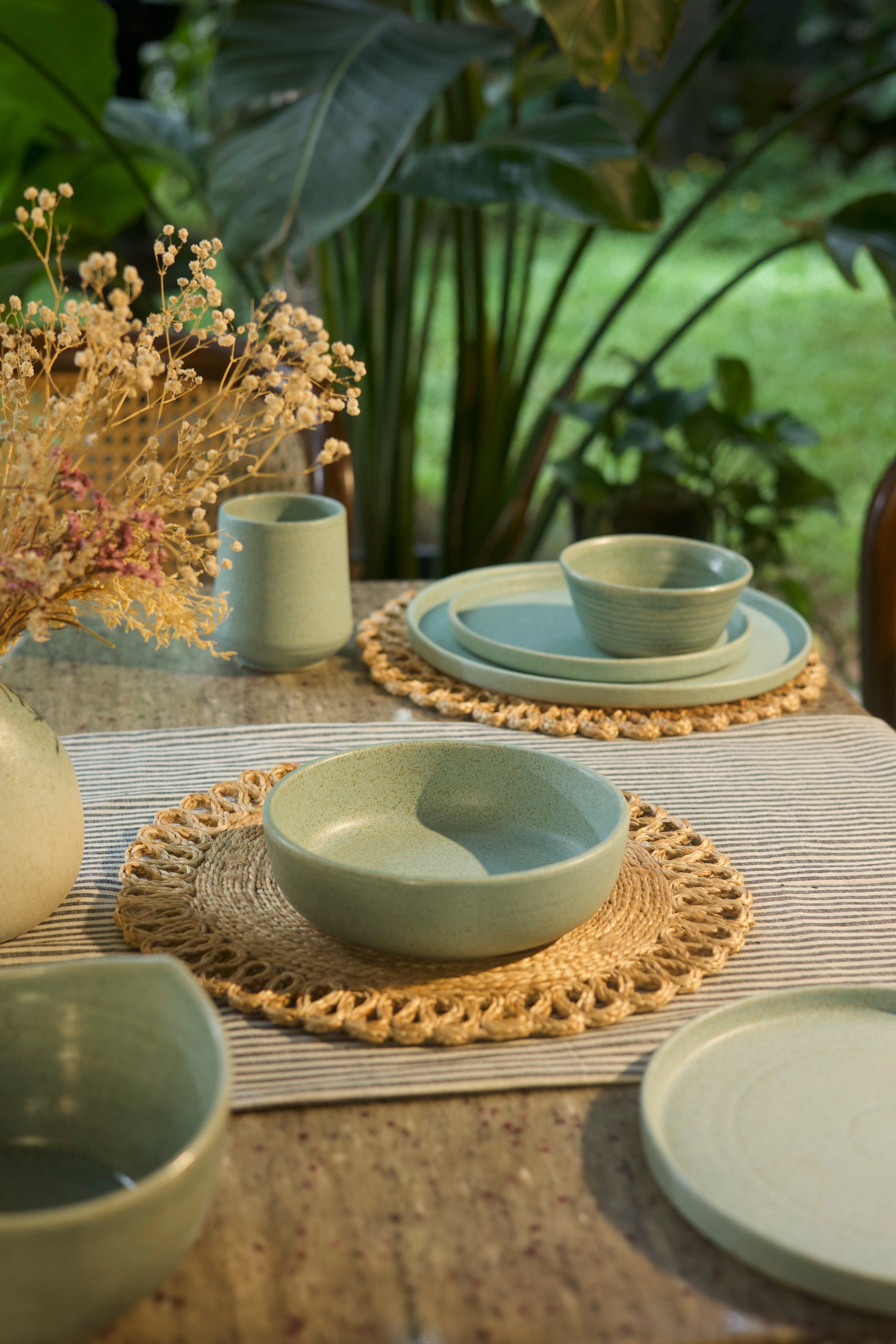 Set of green ceramic dishes on a table with woven placemats and plants in the background, artisanal serveware India, handcrafted plates and bowls