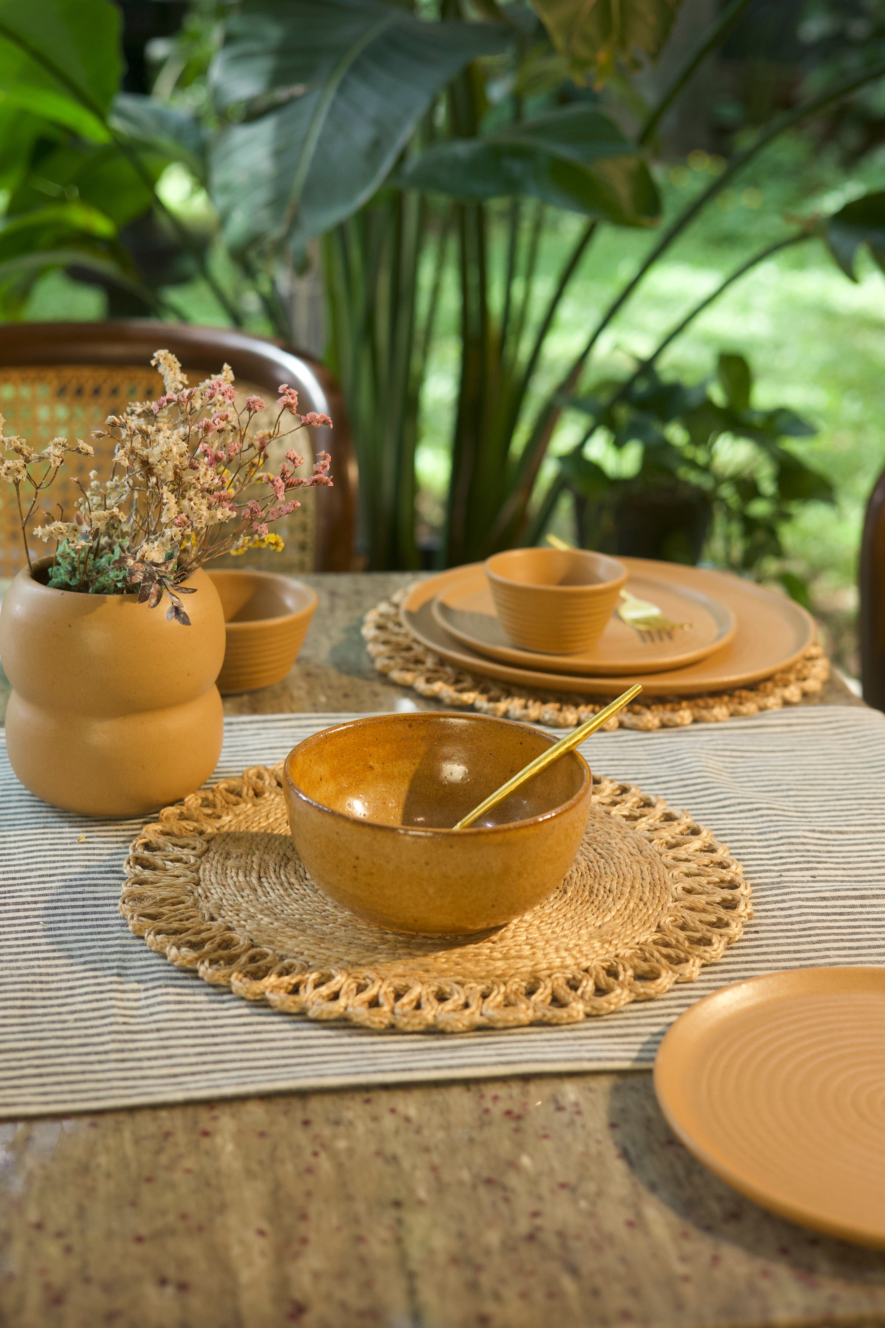 Mustard ceramic bowls and plates on a table with woven placemats, surrounded by plants. artisanal serveware India, handcrafted plates and bowls