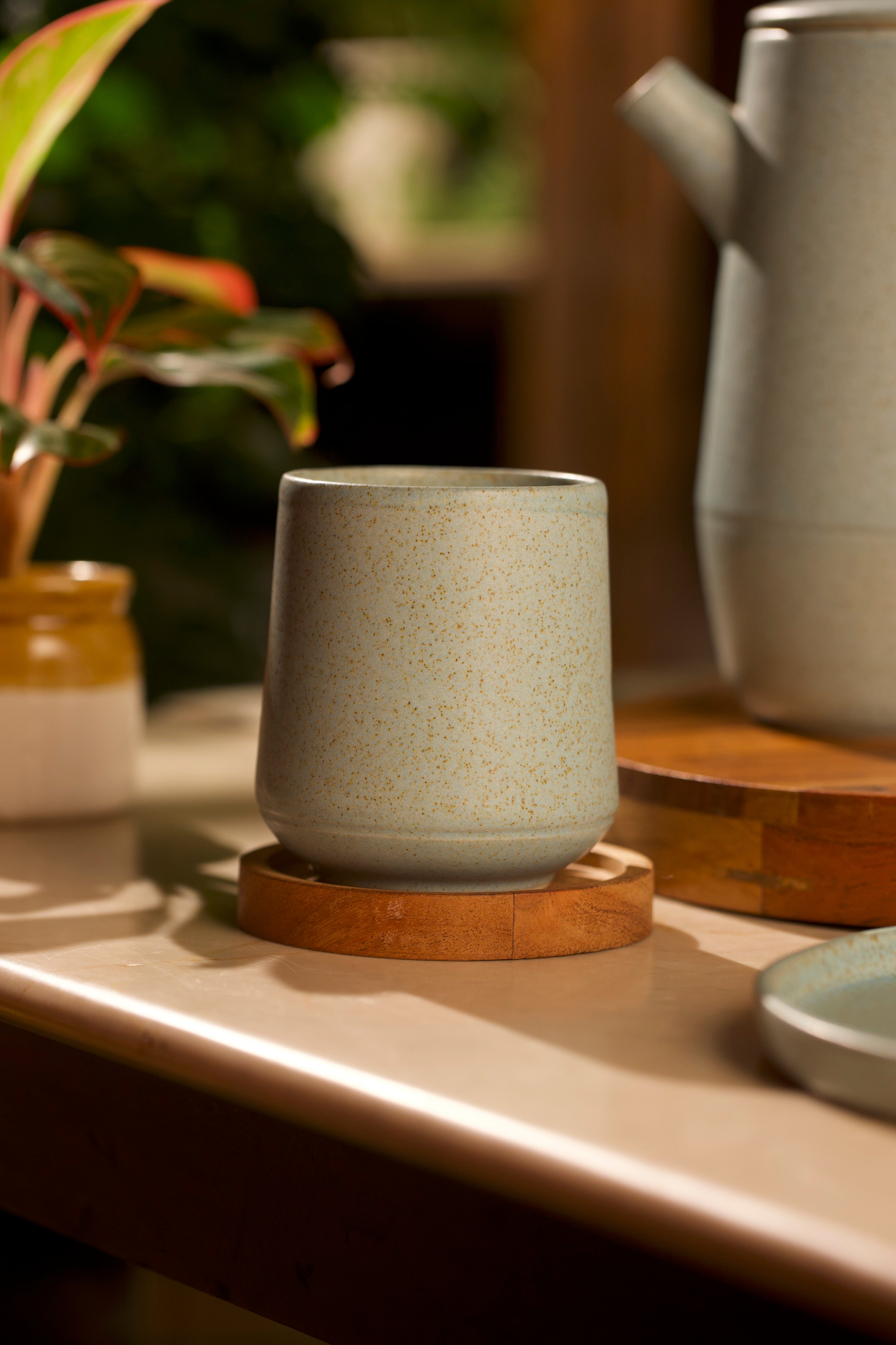 Ceramic cup on a wooden coaster with a blurred background of plants and kitchen items. artisanal serveware India, handmade ceramics India