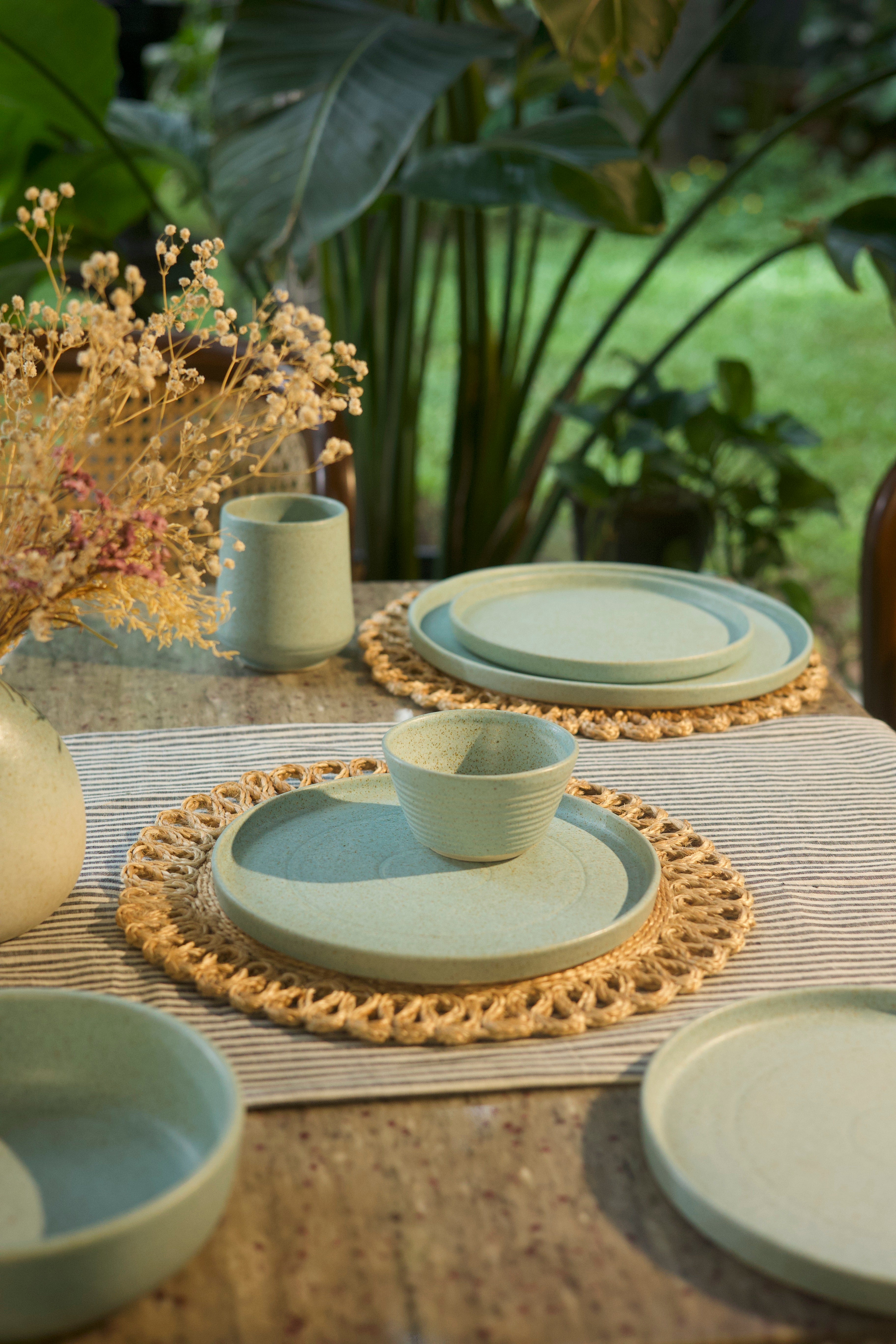 Set of ceramic plates and cups on a table with woven placemats and plants in the background handmade dinnerware India, handcrafted plates and bowls