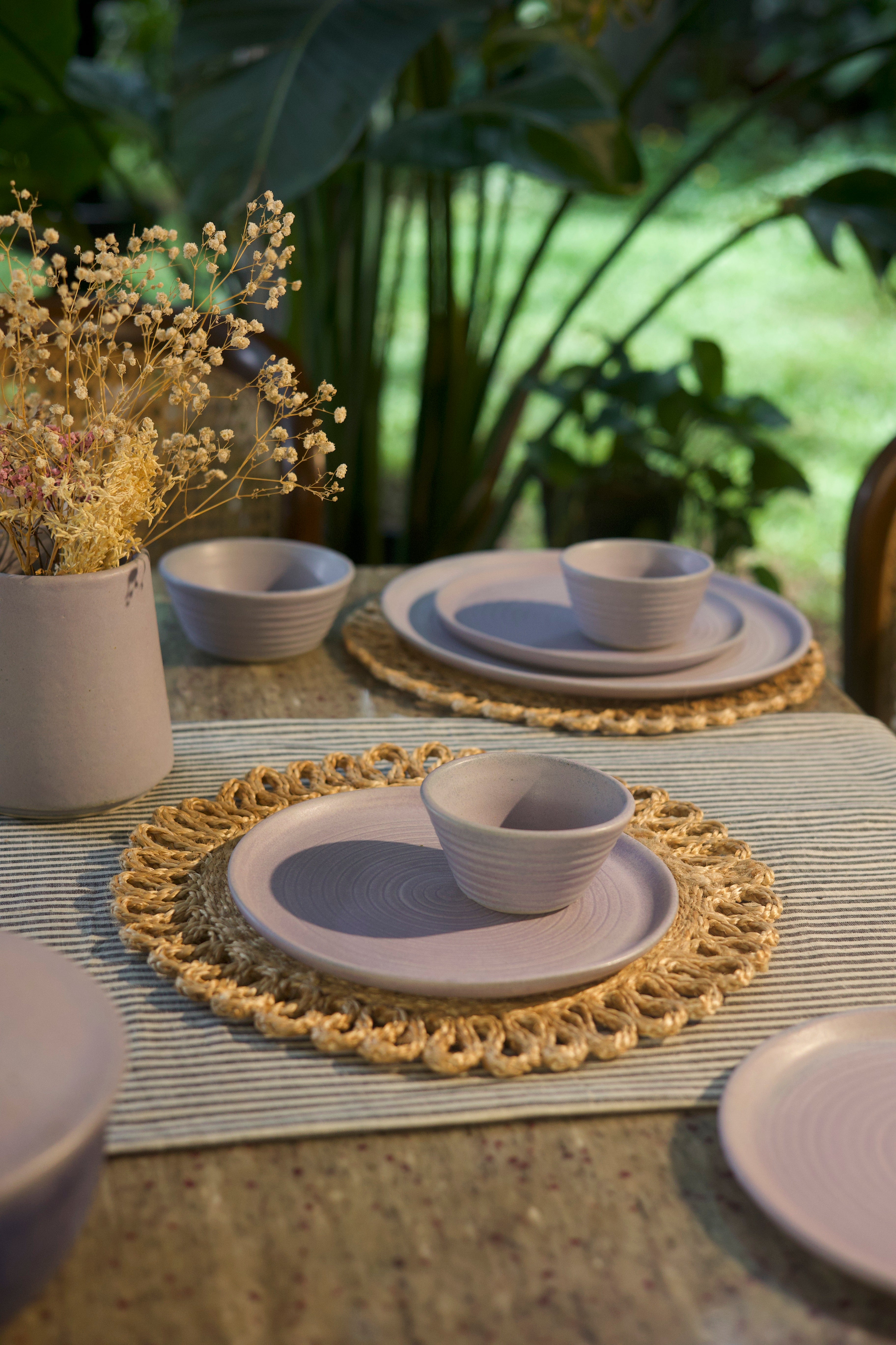 Dining table setting with ceramic plates and bowls on a textured surface with plants in the background. handmade dinnerware India, handcrafted plates and bowls
