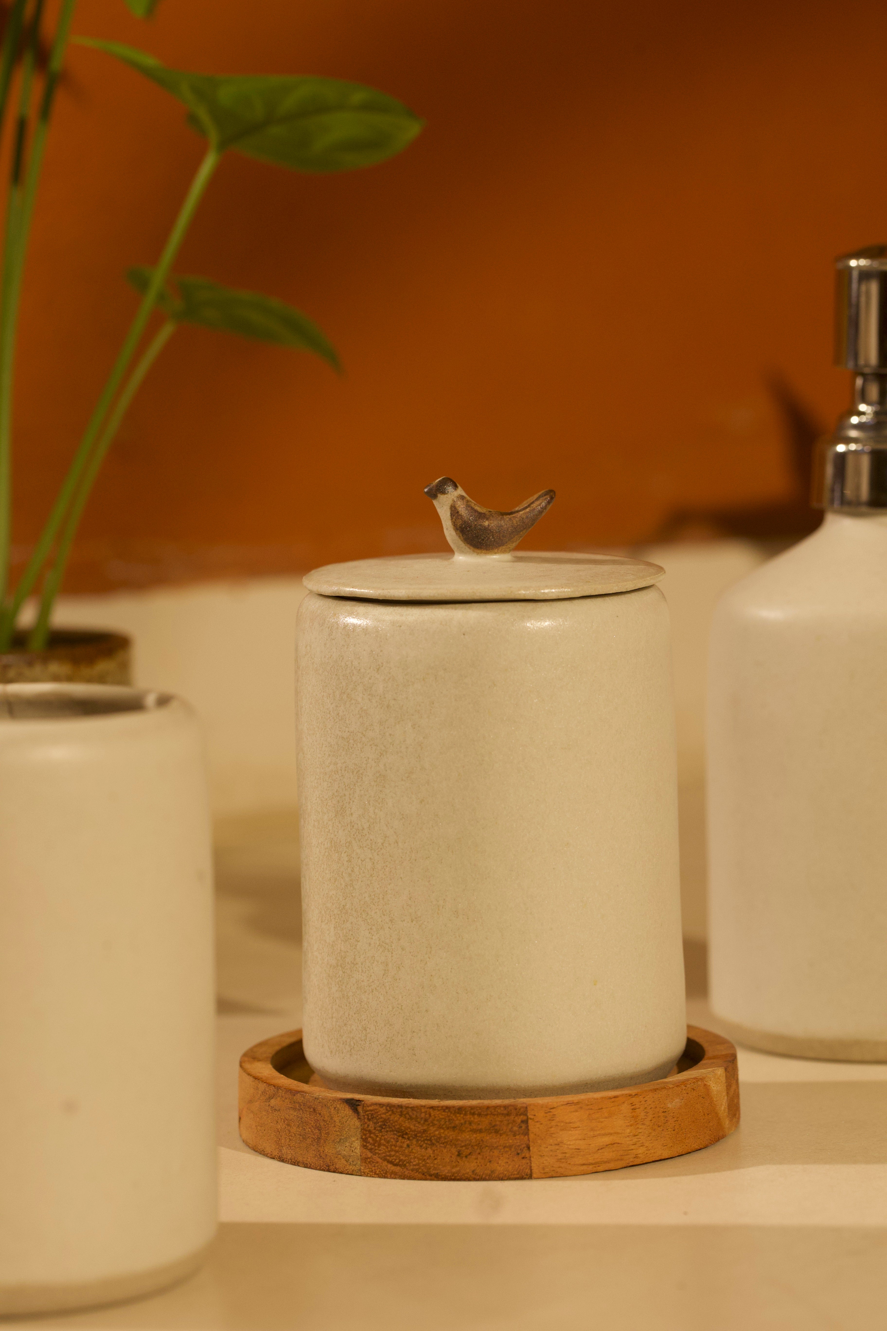 Ceramic jar with bird design on lid on a wooden coaster, surrounded by plants and other ceramic items. ceramic home décor online, pottery home decor