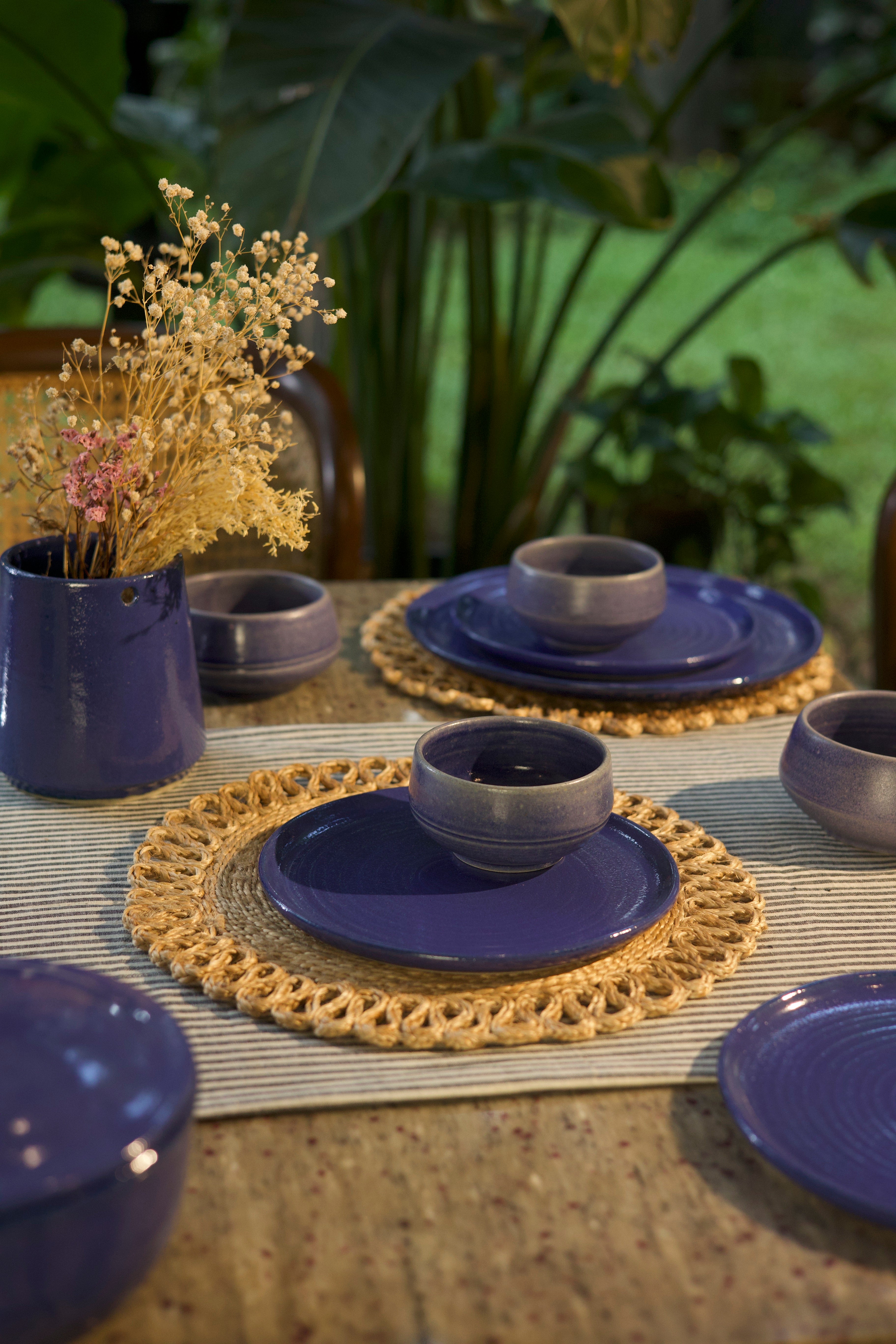 Set of blue ceramic plates and bowls on a table with woven placemats and a plant in the background. handmade dinnerware India, handcrafted plates and bowls