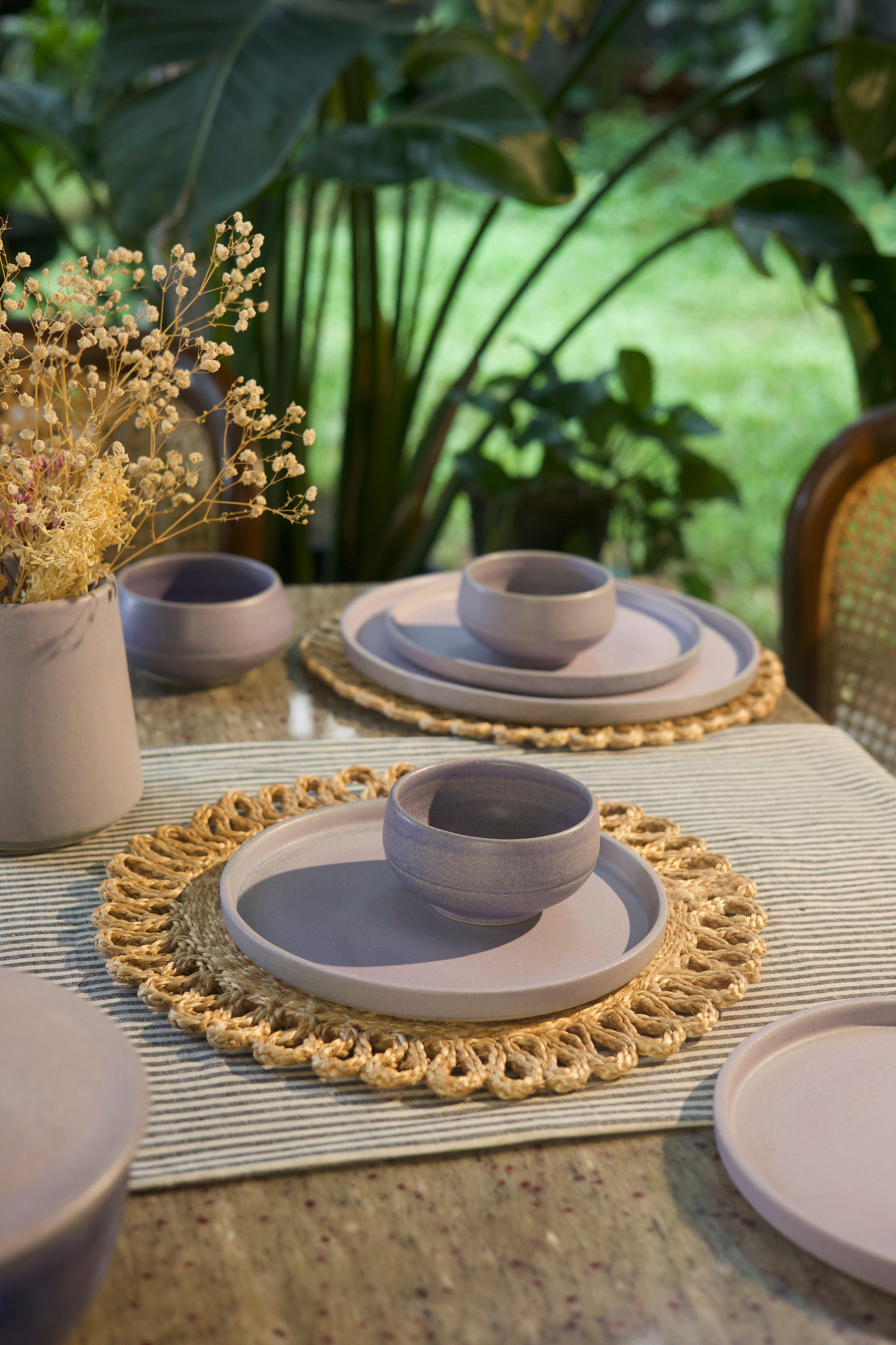 Dining table setting with ceramic plates and bowls on a textured surface with greenery in the background, handmade dinnerware India, handcrafted plates and bowls