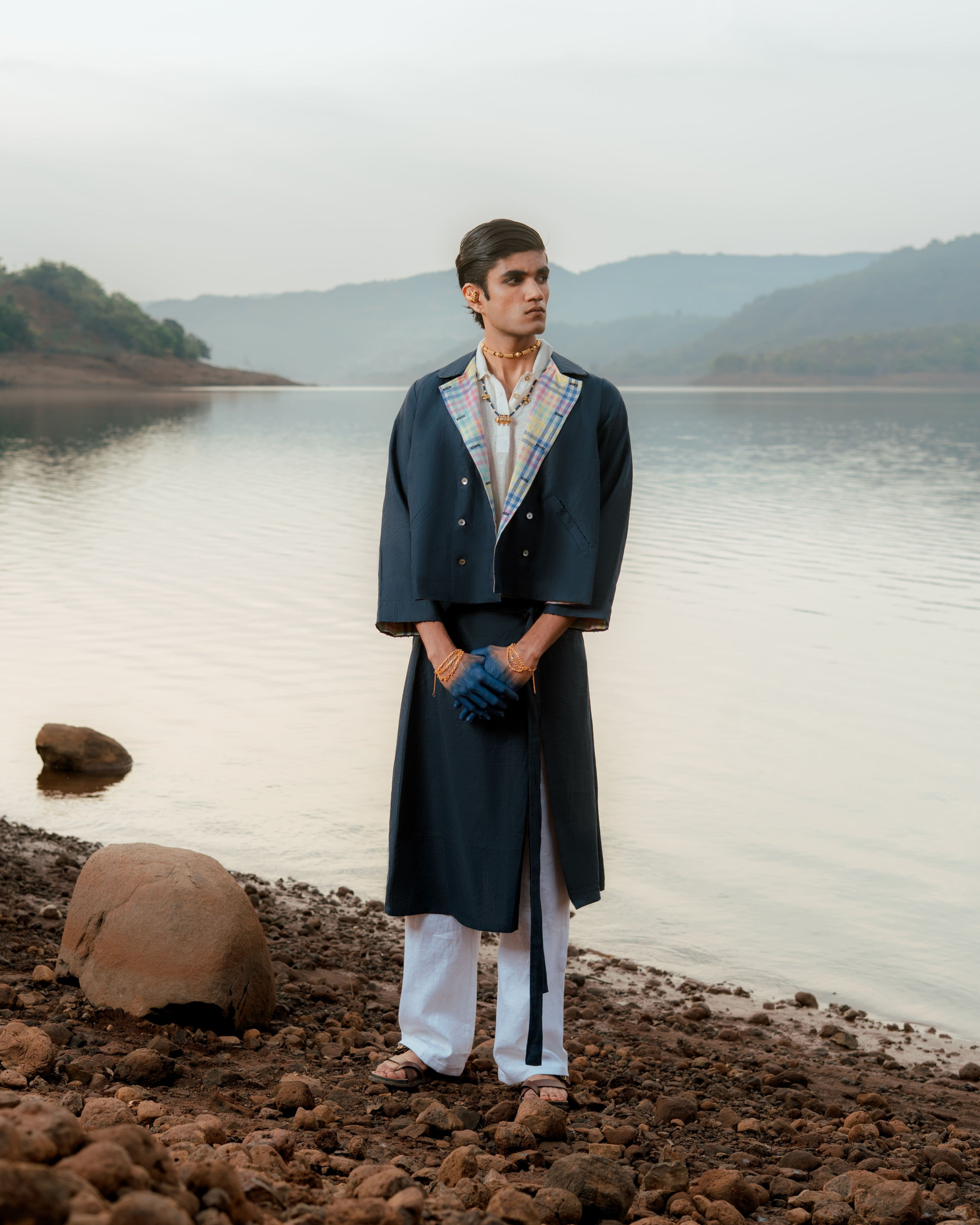 Man standing by a lake wearing a dark coat and white pants,  sustainable fashion brands for men,
linen shirts for men India
