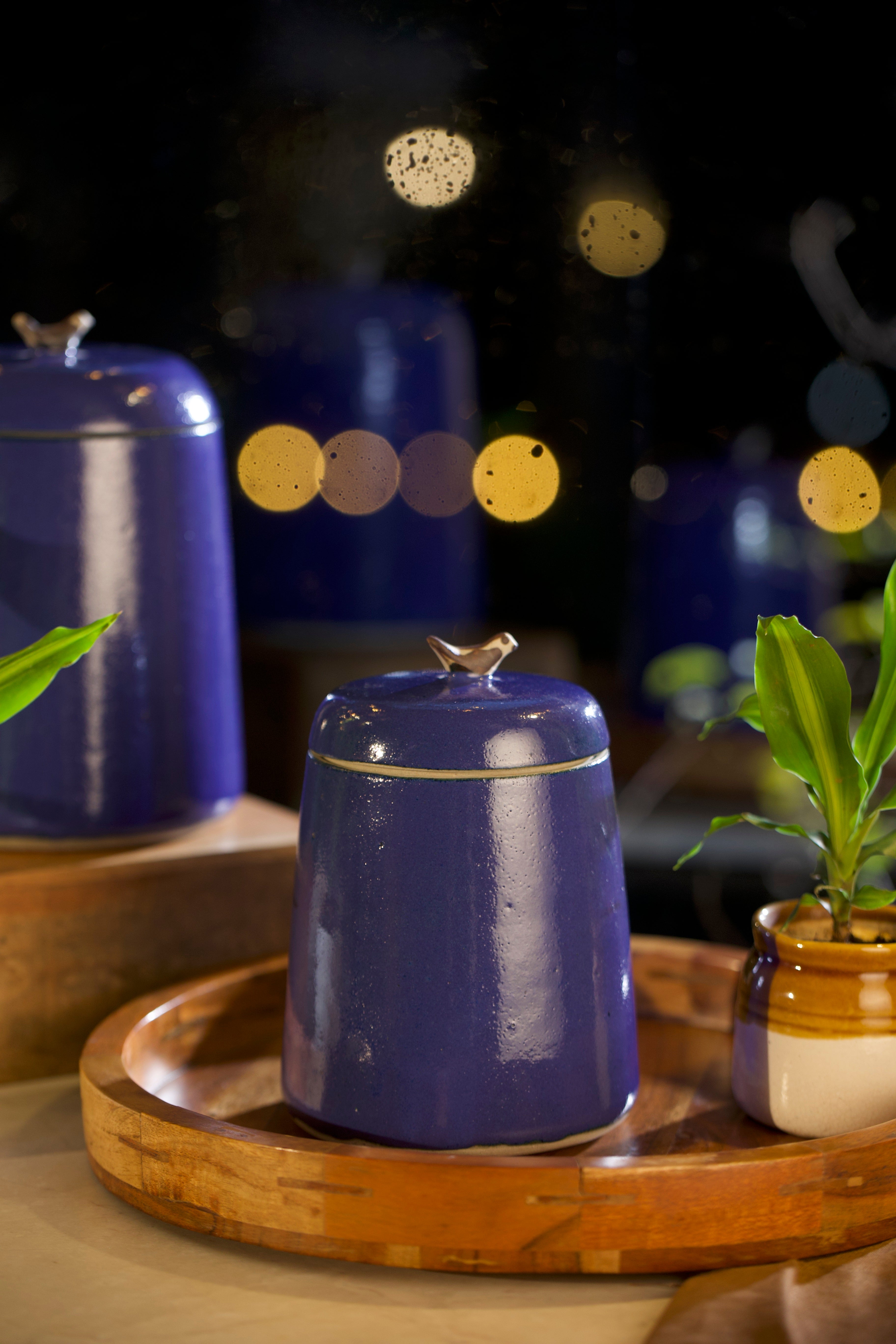 Blue ceramic jar with a lid on a wooden tray with blurred lights in the background,  pottery home decor, handmade serveware online
