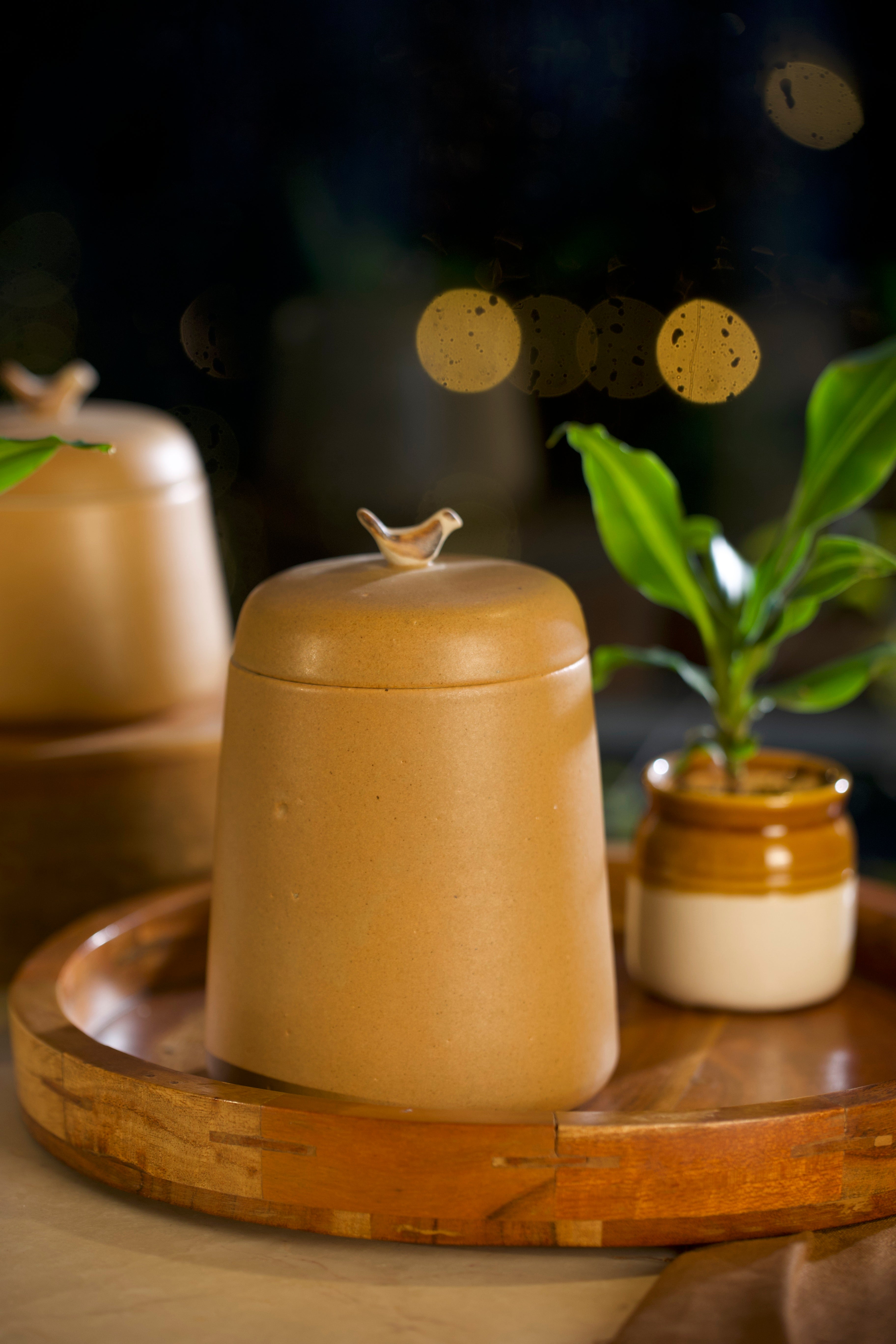 A mustard ceramic jar with a lid featuring a small bird ornament on a wooden tray. pottery home decor, handmade serveware online