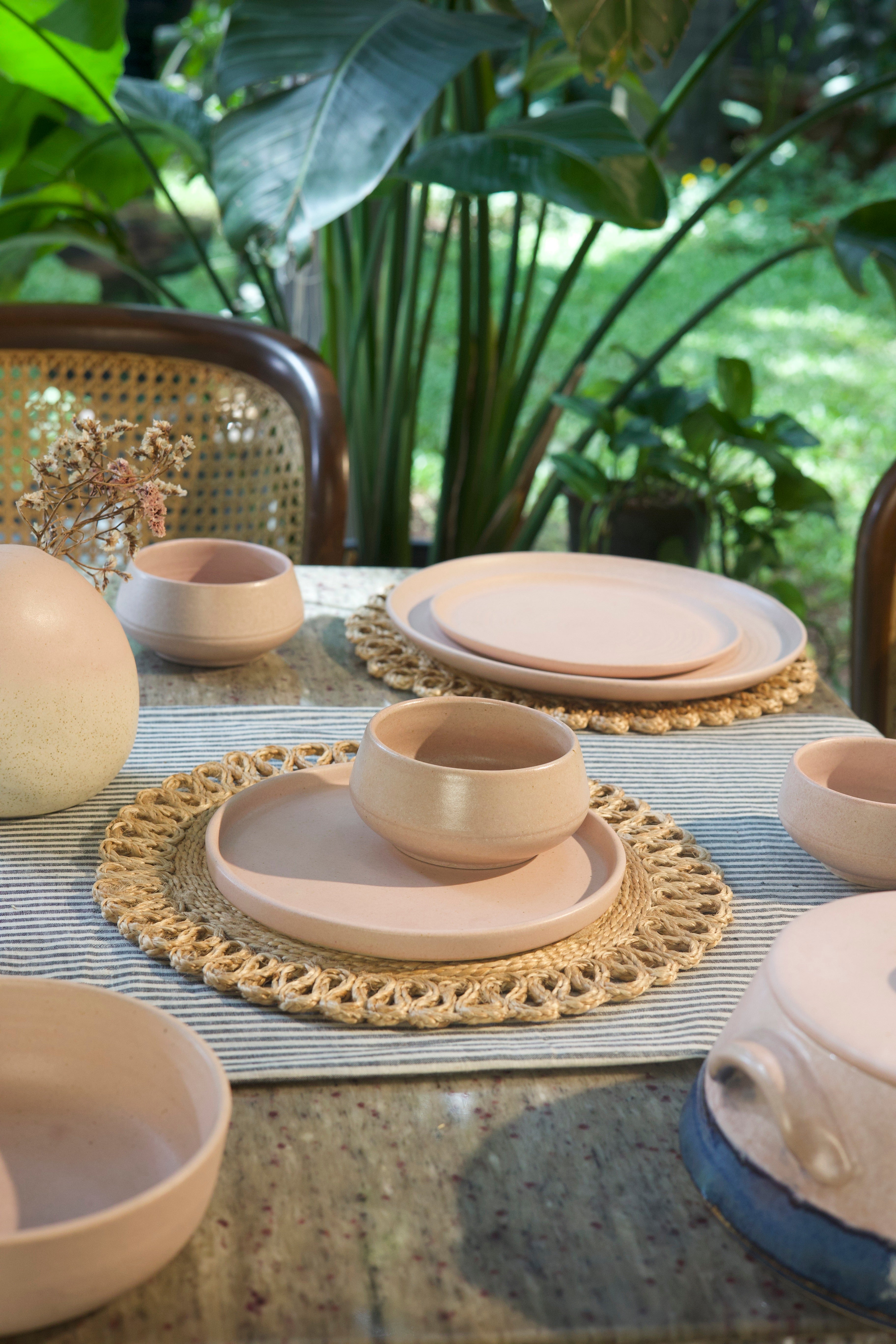 Table setting with ceramic plates and bowls on a textured surface with plants in the background, handmade dinnerware India, handcrafted plates and bowls