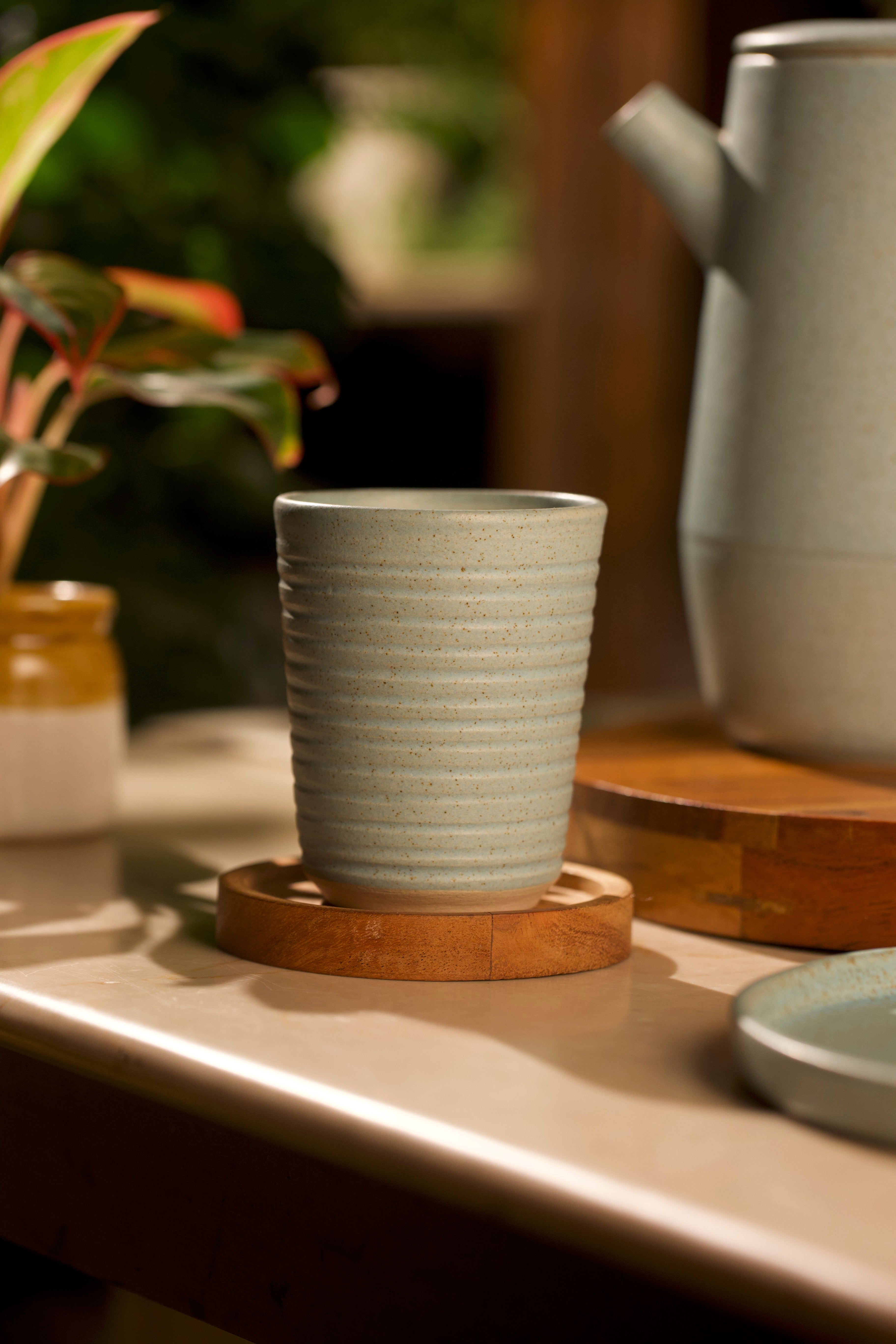 Ceramic cup on a wooden coaster with a teapot and plant in the background, artisanal serveware India, handmade ceramics India 
